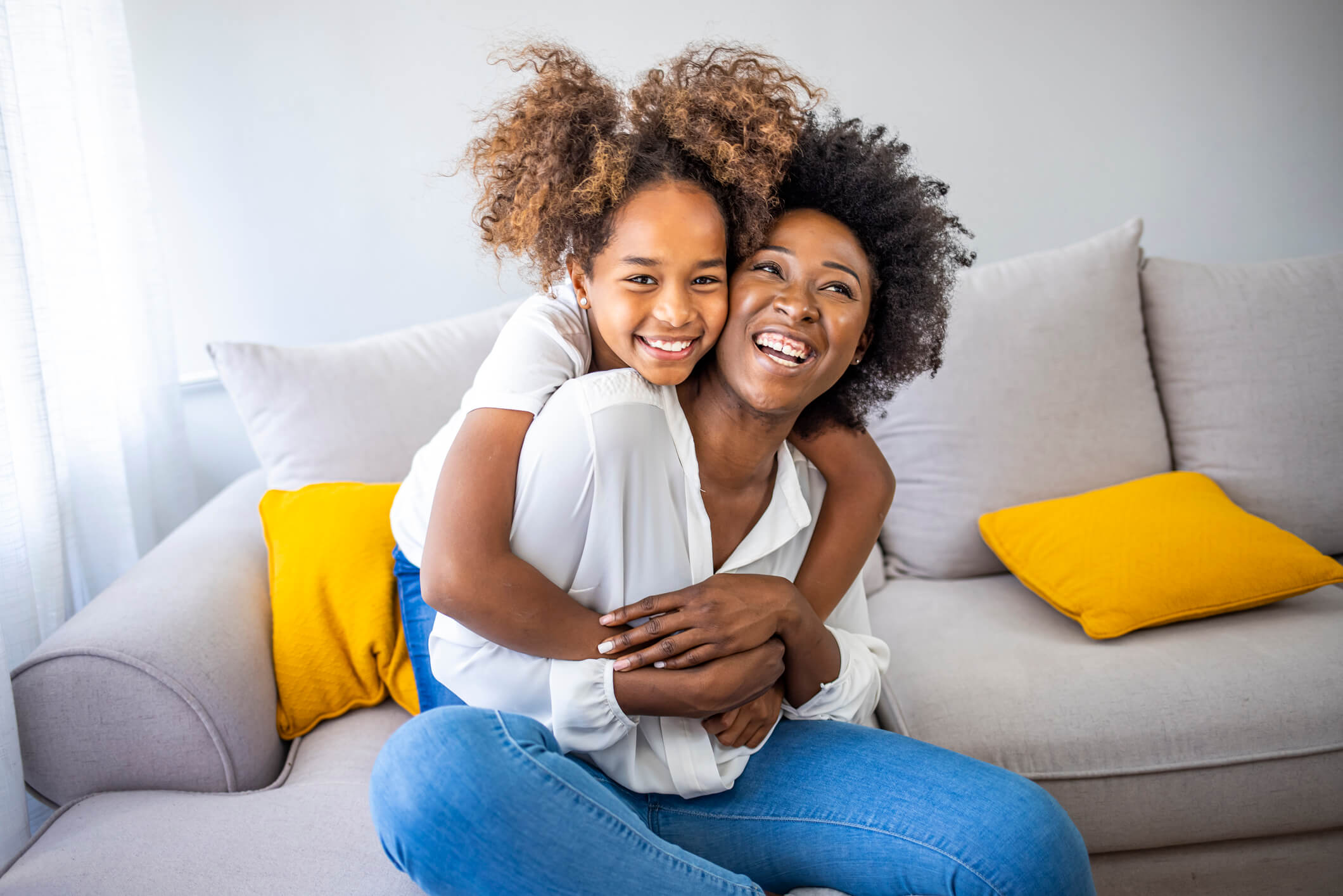 Mother sitting down on a couch with her young daughter hugging her from behind.