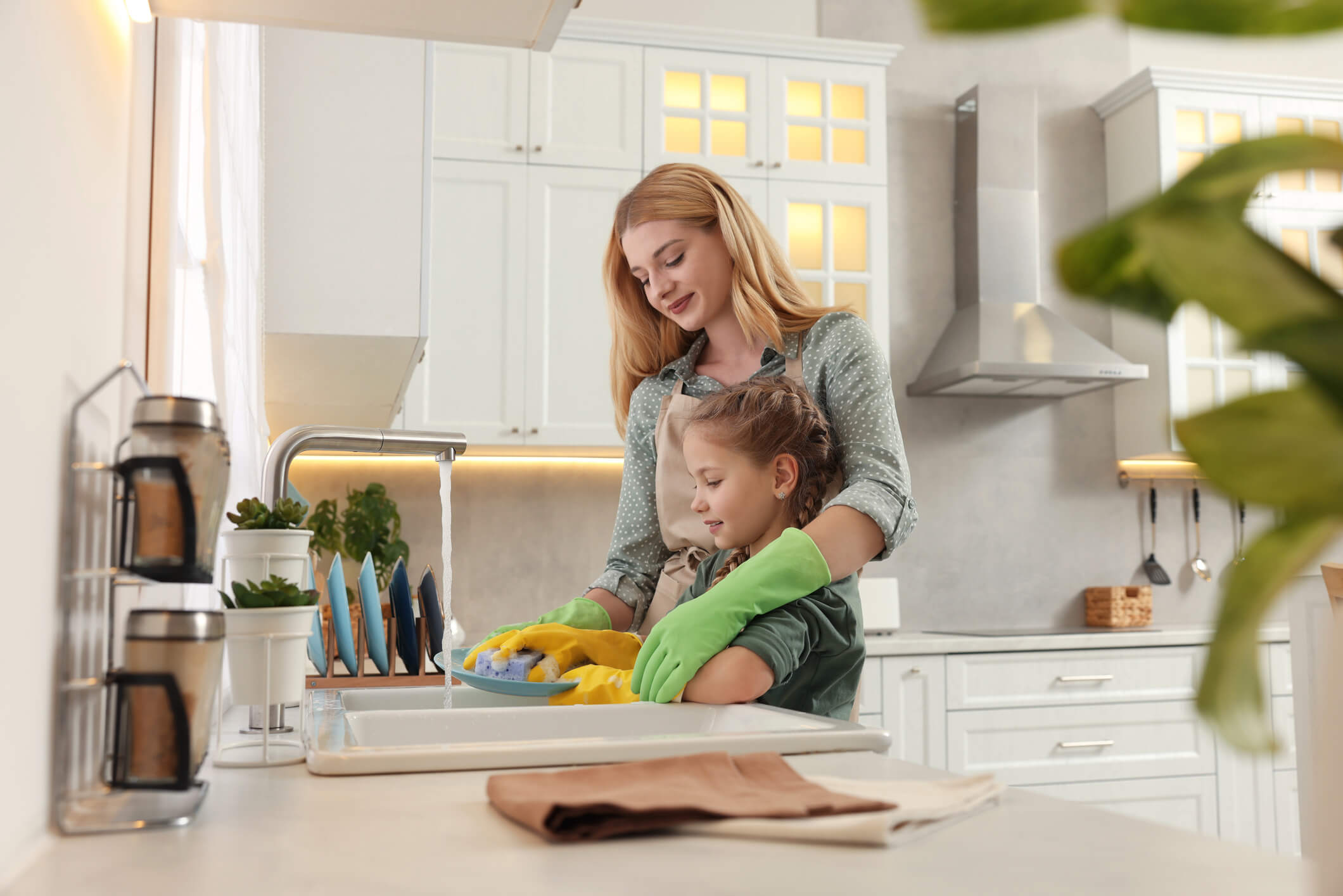 Mom wearing green dishwashing gloves helping her son, wearing yellow dishwashing gloves, wash dishes in the kitchen sink.