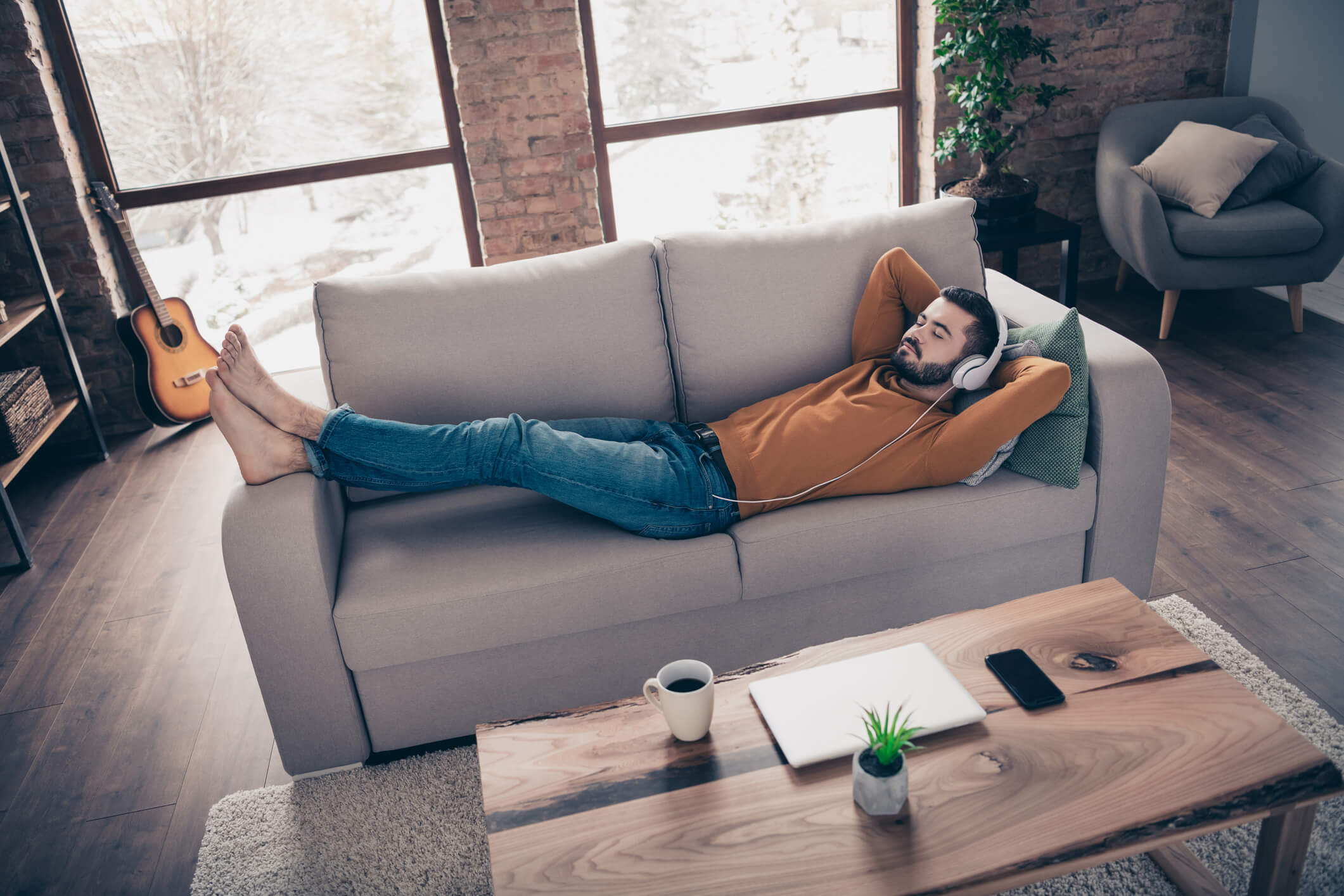 Young man wearing a yellow sweater and jeans laying on the couch while wearing headphones and listening to music.