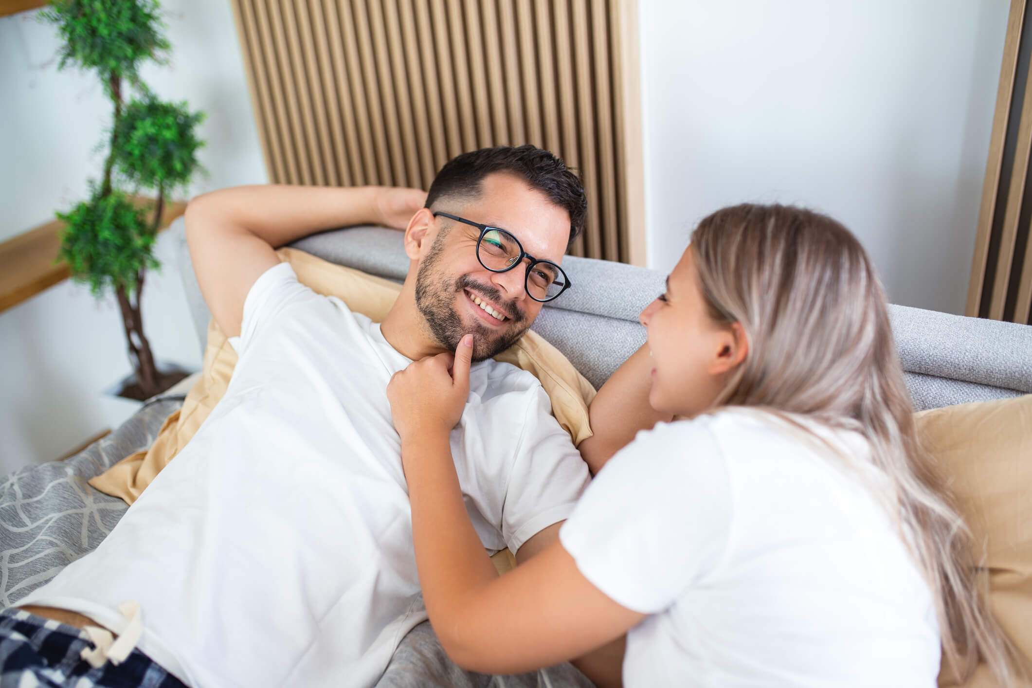 Young couple laying in bed. Woman in the couple is holding the chin of her male partner.