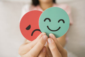 Image of a person's hands holding a sad face card hiding behind a happy face card, symbolizing bipolar disorder and depression in mental health.