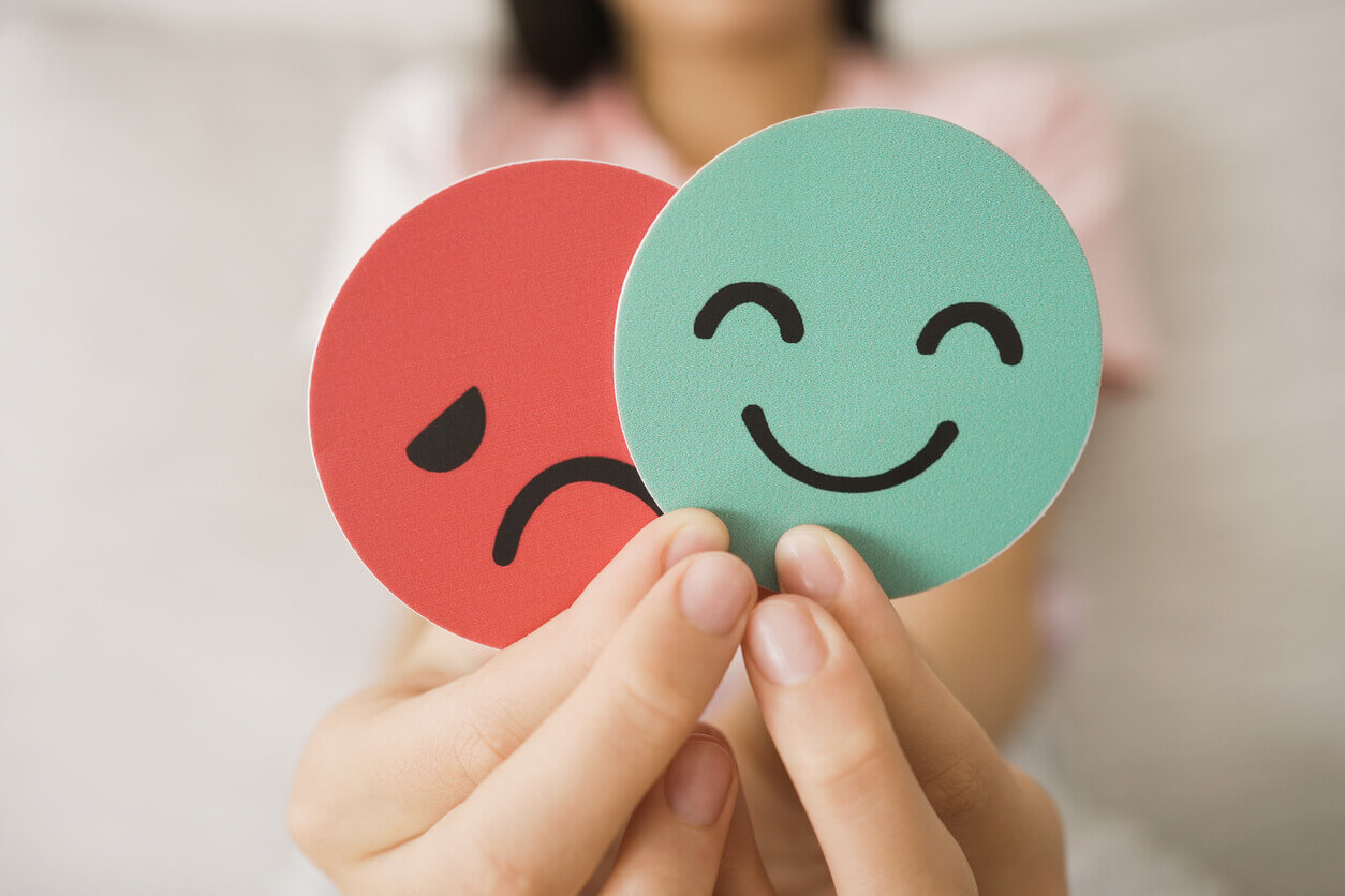Image of a person's hands holding a sad face card hiding behind a happy face card, symbolizing bipolar disorder and depression in mental health.