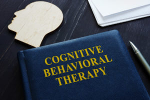 Cognitive Behavioral Therapy (CBT) is written on a book placed on a table alongside a pen and a wooden head shape.