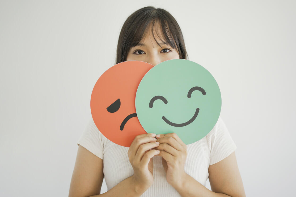 Woman hands holding sad face hiding behind happy face due to bipolar mood disorder.