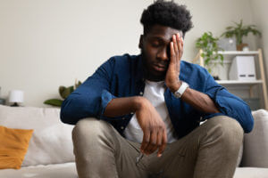 Overwhelmed African American man sitting on the couch, having anxiety and feeling sad.